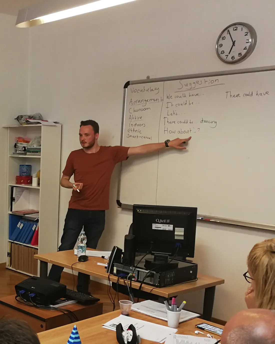 Anthony at a whiteboard, pointing to a list of phrases for making suggestions, during a lesson